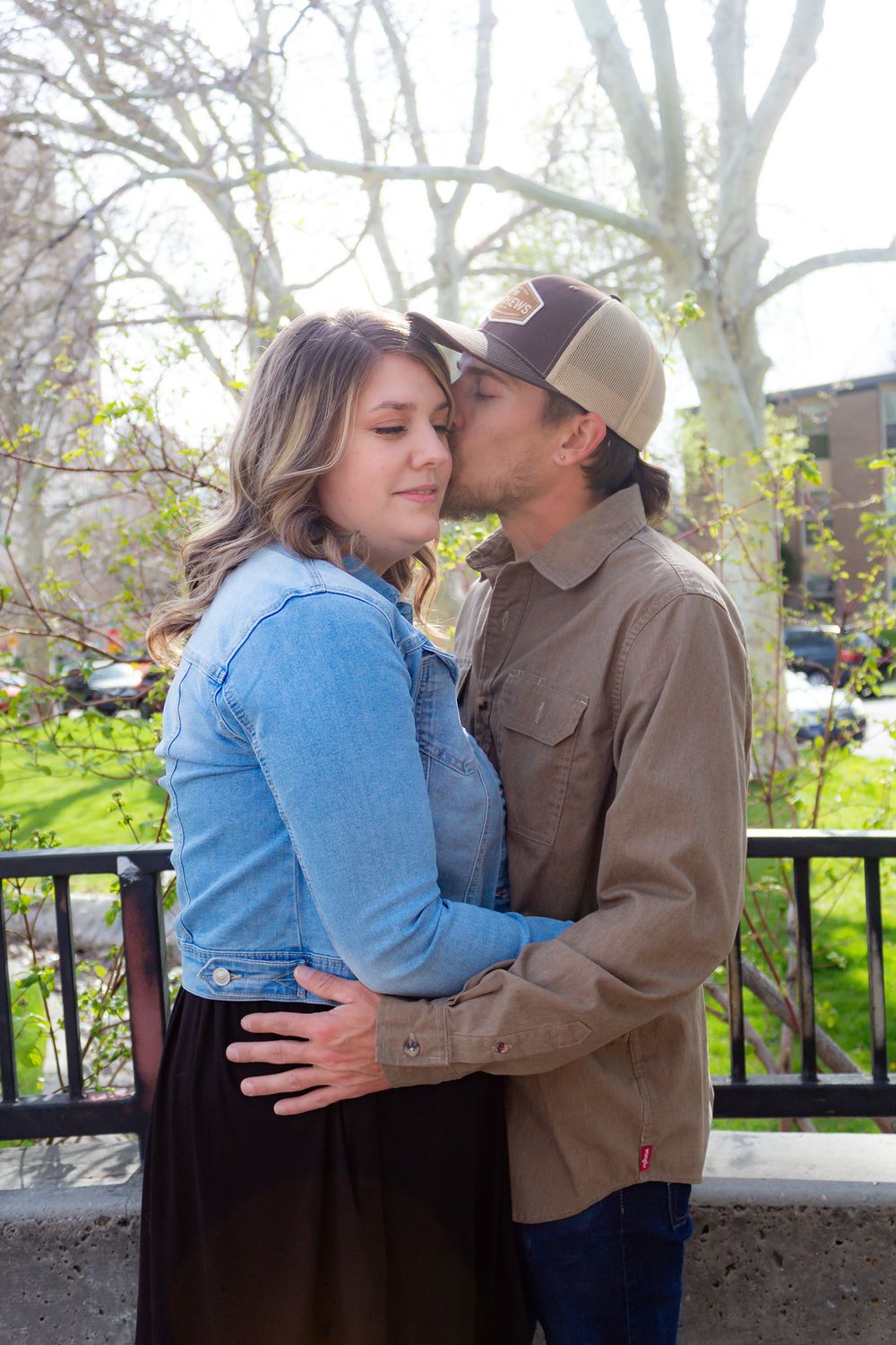 A romantic couple embraces outdoors in spring, with her in a blue jacket and him in brown clothing and baseball cap.
