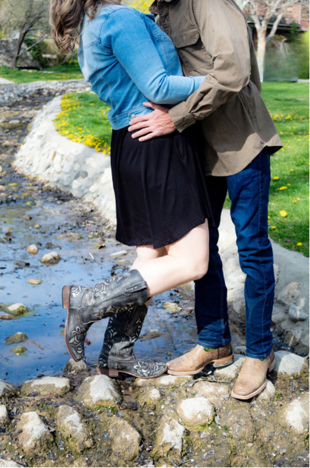 Couple stands on rocky terrain near water, with her wearing cowboy boots and him in casual western wear.