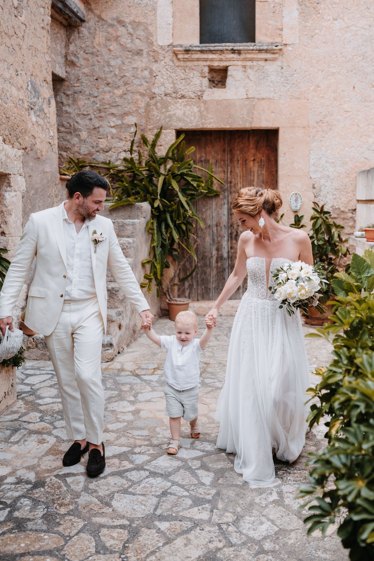 Ein Paar in Hochzeitskleidung spaziert mit einem kleinen Kind zwischen sich auf Kopfsteinpflaster in einer rustikalen mediterranen Location.