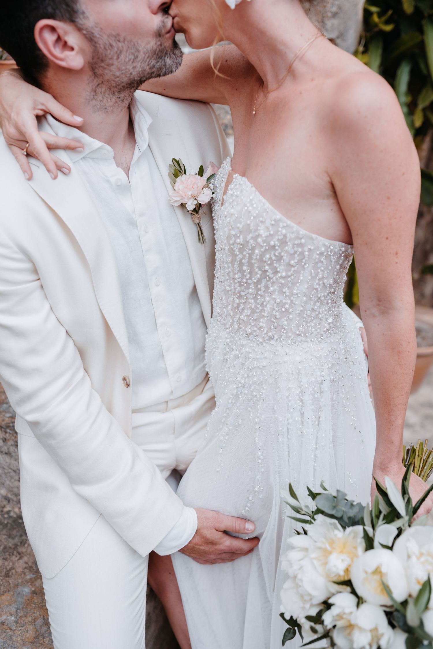 Romantisches Detailbild, das ein Brautpaar in weißer Kleidung mit Blumenanstecksträußchen und funkelnden Details am Brautkleid zeigt.