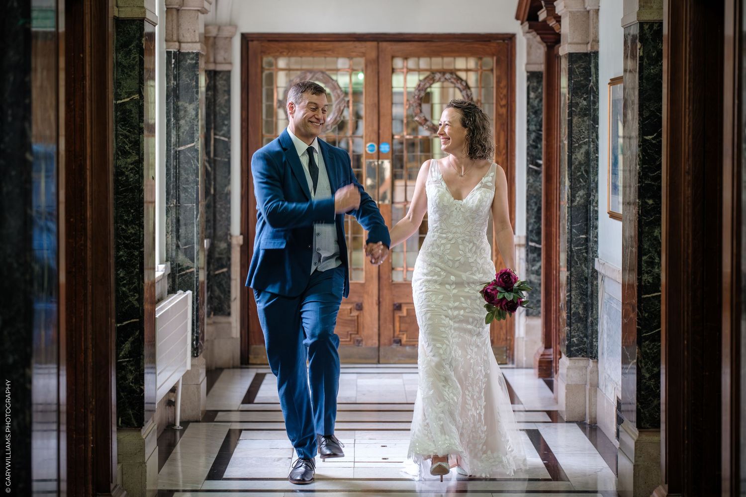 A couple walks down an elegant marble hallway on their wedding day, with ornate wooden doors in the background.