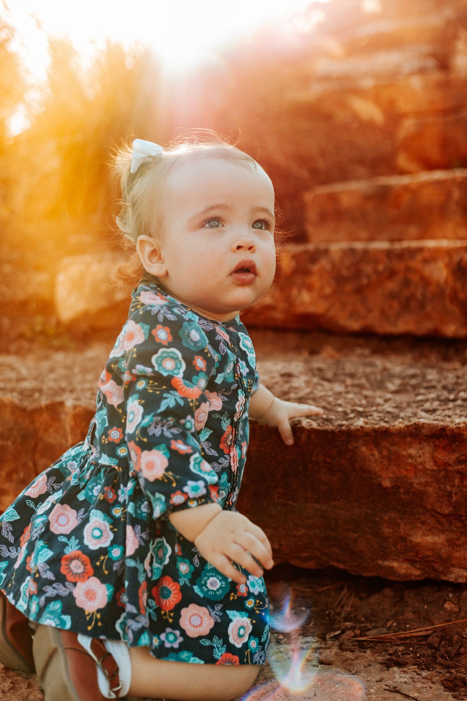 Toddler girl in floral dress sitting by stone steps in golden sunset light outdoors.