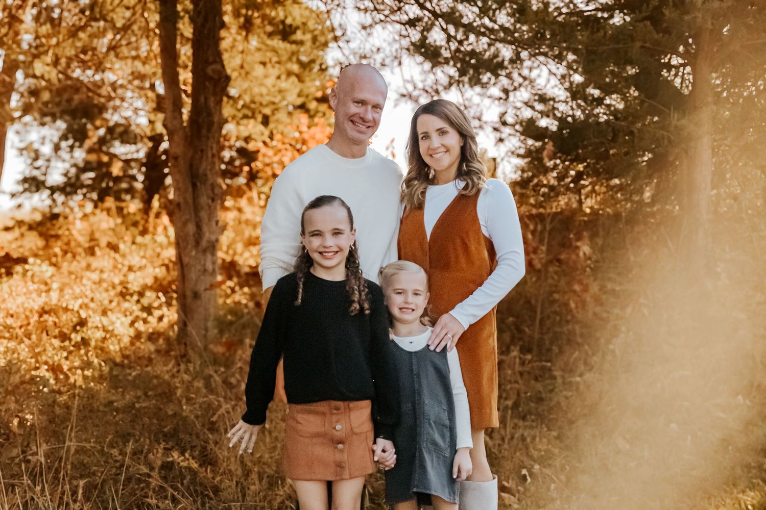 Family of four posing outdoors in autumn, parents and two daughters smiling among golden fall foliage.