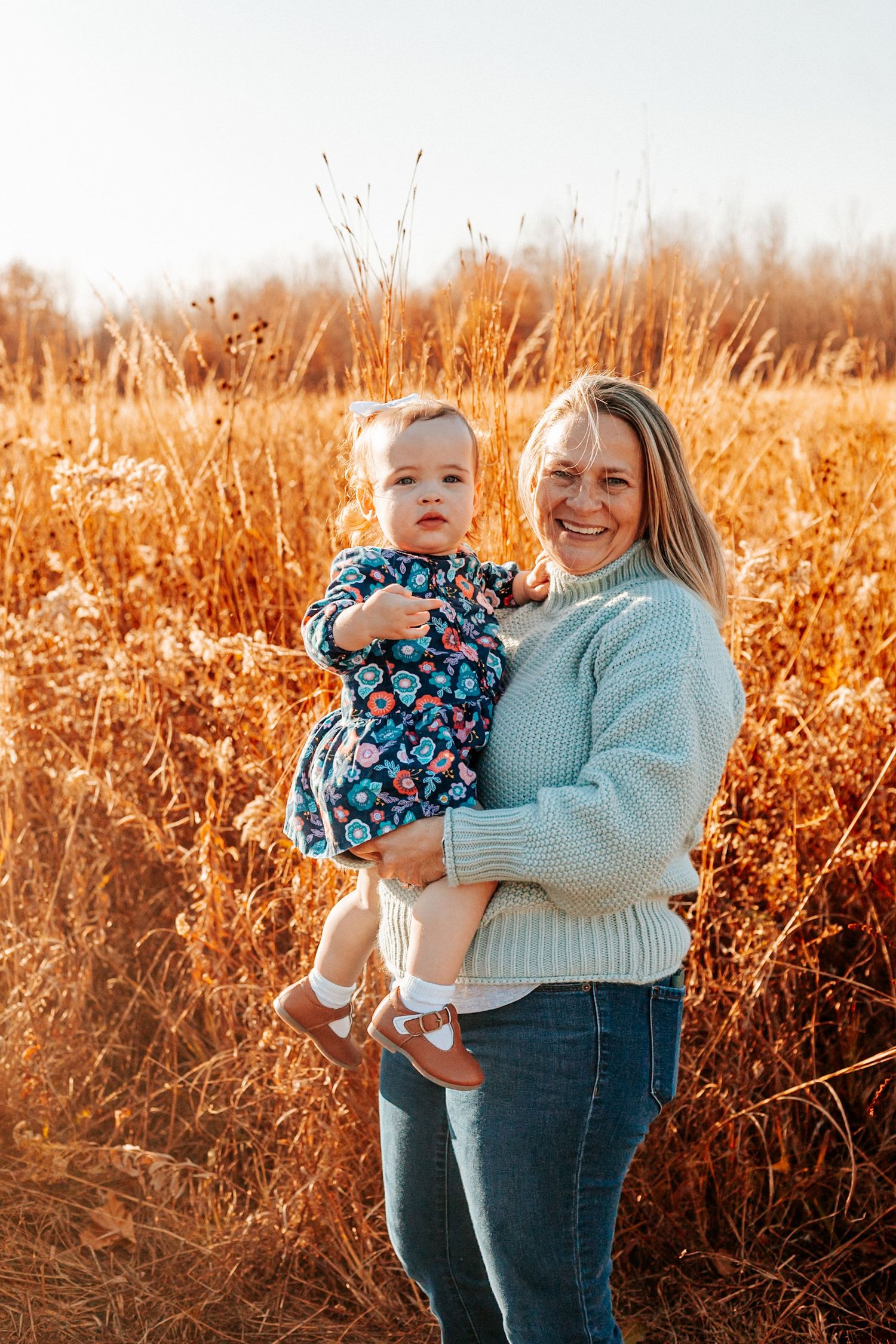 A smiling woman in a light blue sweater holds a baby in a floral dress in a golden autumn field.