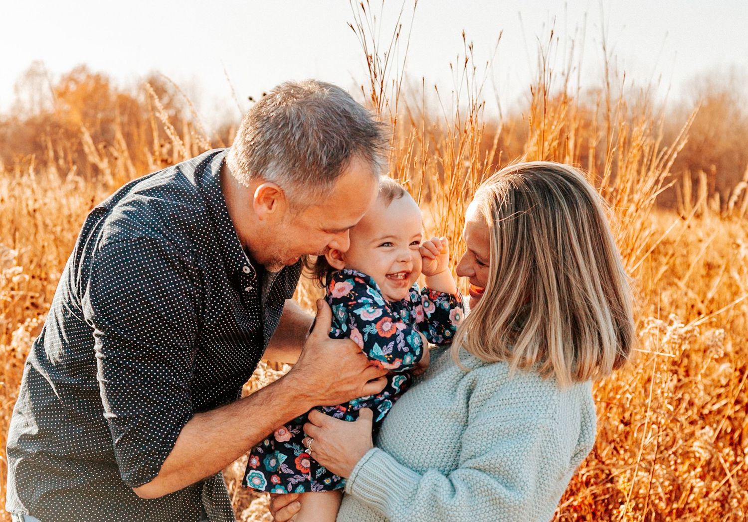 Happy family with parents kissing smiling baby girl in sunny autumn field with golden tall grass.