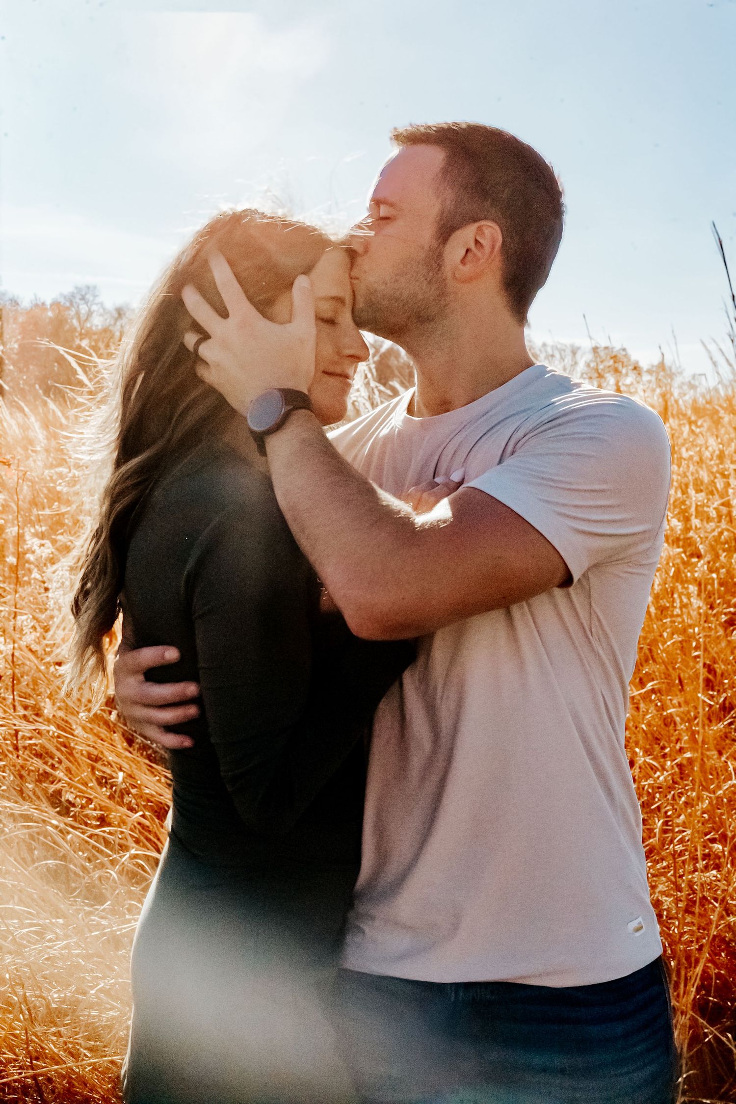 A couple shares a tender kiss in a sunlit golden wheat field, with the man gently holding the woman's face.