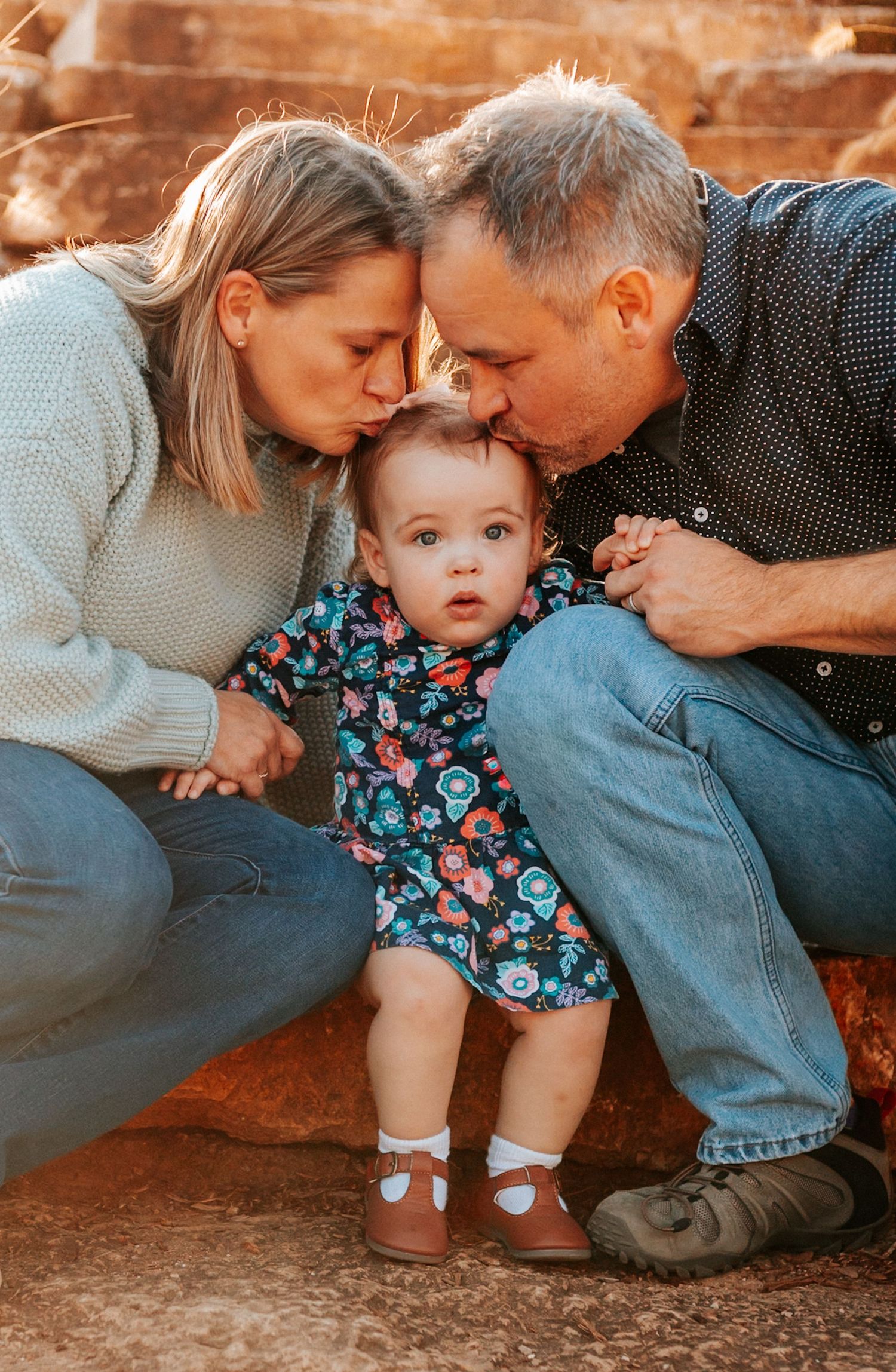 Parents kissing baby girl on cheeks during outdoor family portrait session with warm natural lighting.