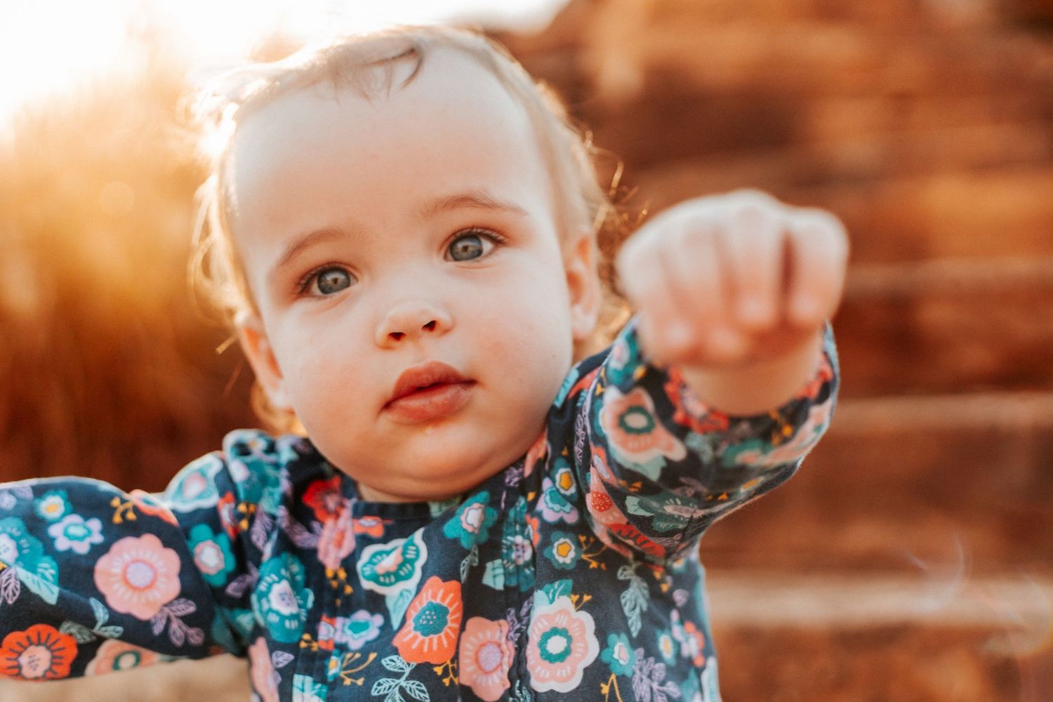 Adorable baby girl in floral outfit pointing finger toward camera in warm golden sunset light outdoors.