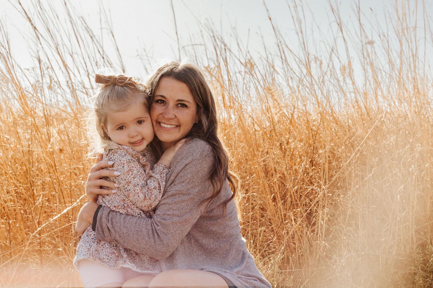 A smiling mother hugs her young daughter in a golden wheat field, both wearing soft neutral tones in warm sunlight.