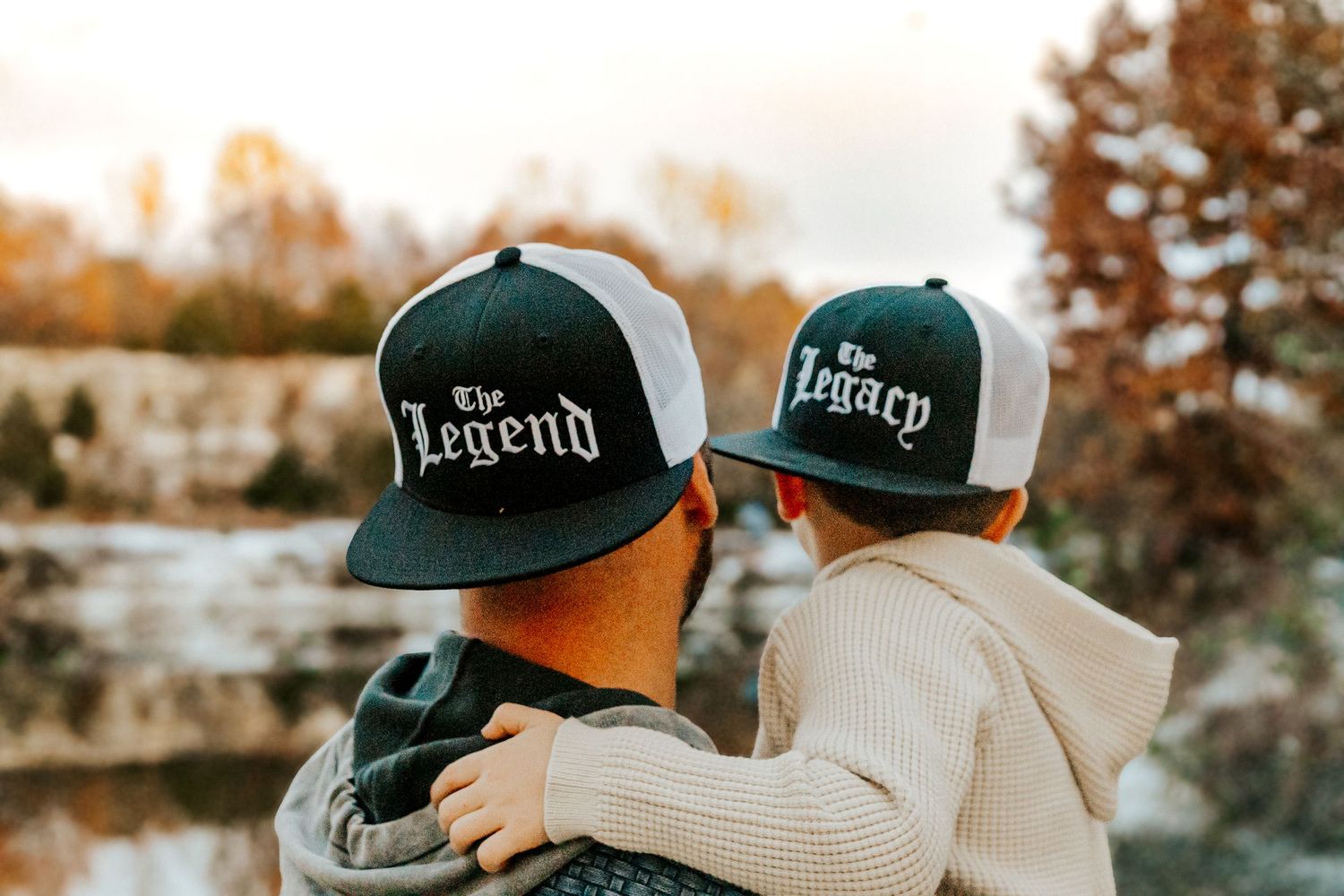 Adult and child wearing matching black trucker hats reading The Legend and The Legacy outdoors near a lake.