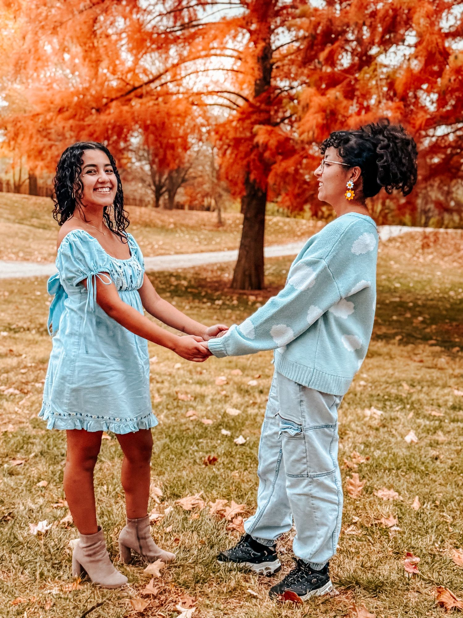 Two women in light blue outfits hold hands in a park surrounded by vibrant autumn foliage.