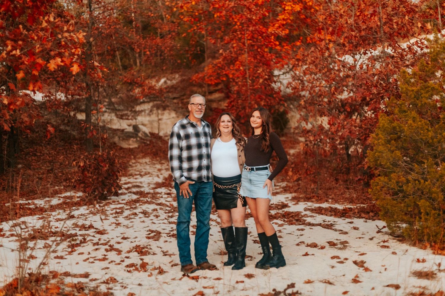 Three people smiling together outdoors among vibrant red autumn foliage on a snowy path.