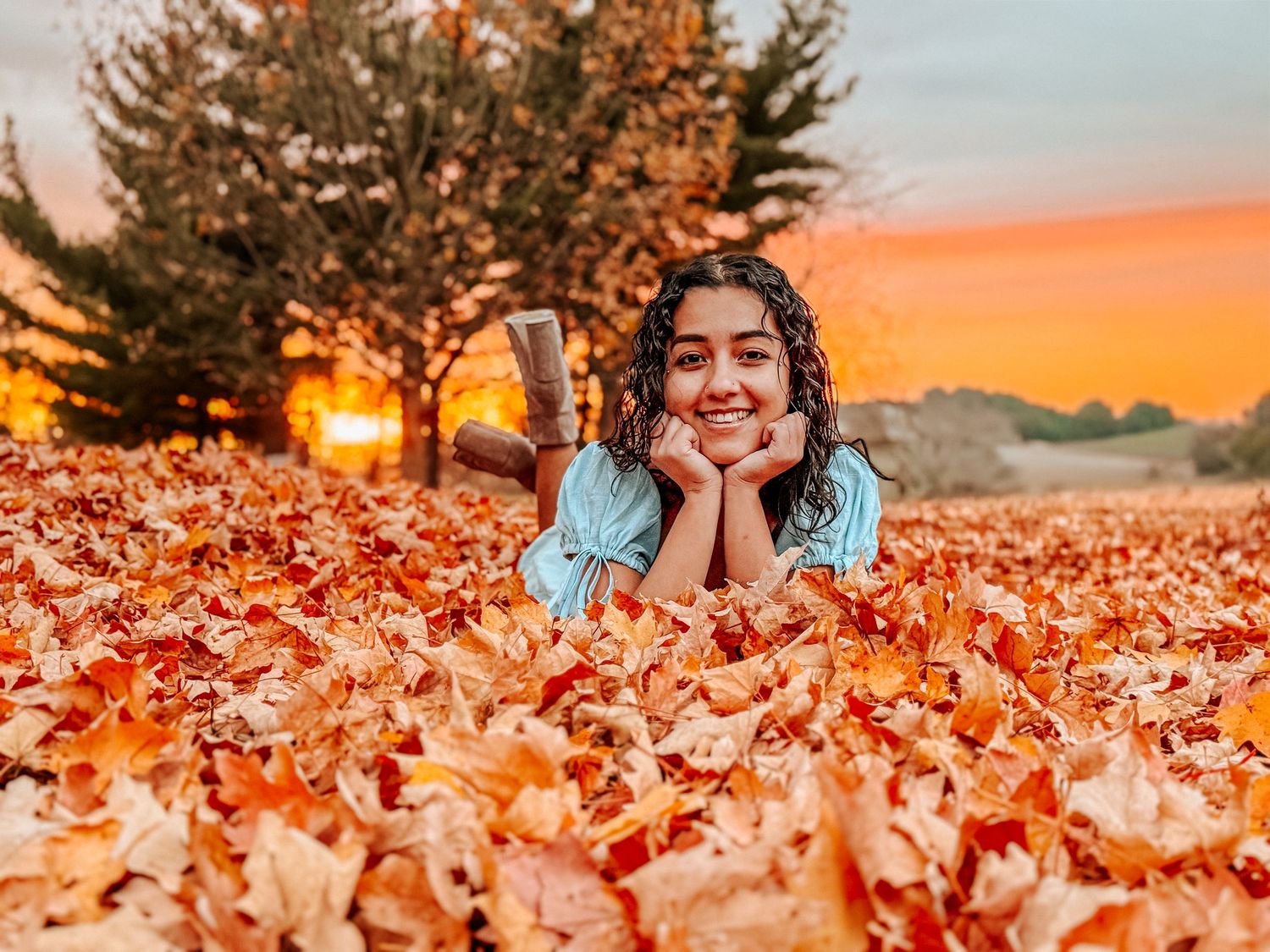 Young woman smiling among vibrant autumn leaves at sunset, lying in a colorful fall foliage landscape.