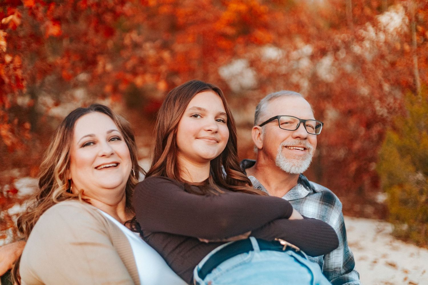 Family of three laughing and embracing outdoors with colorful fall foliage in background.
