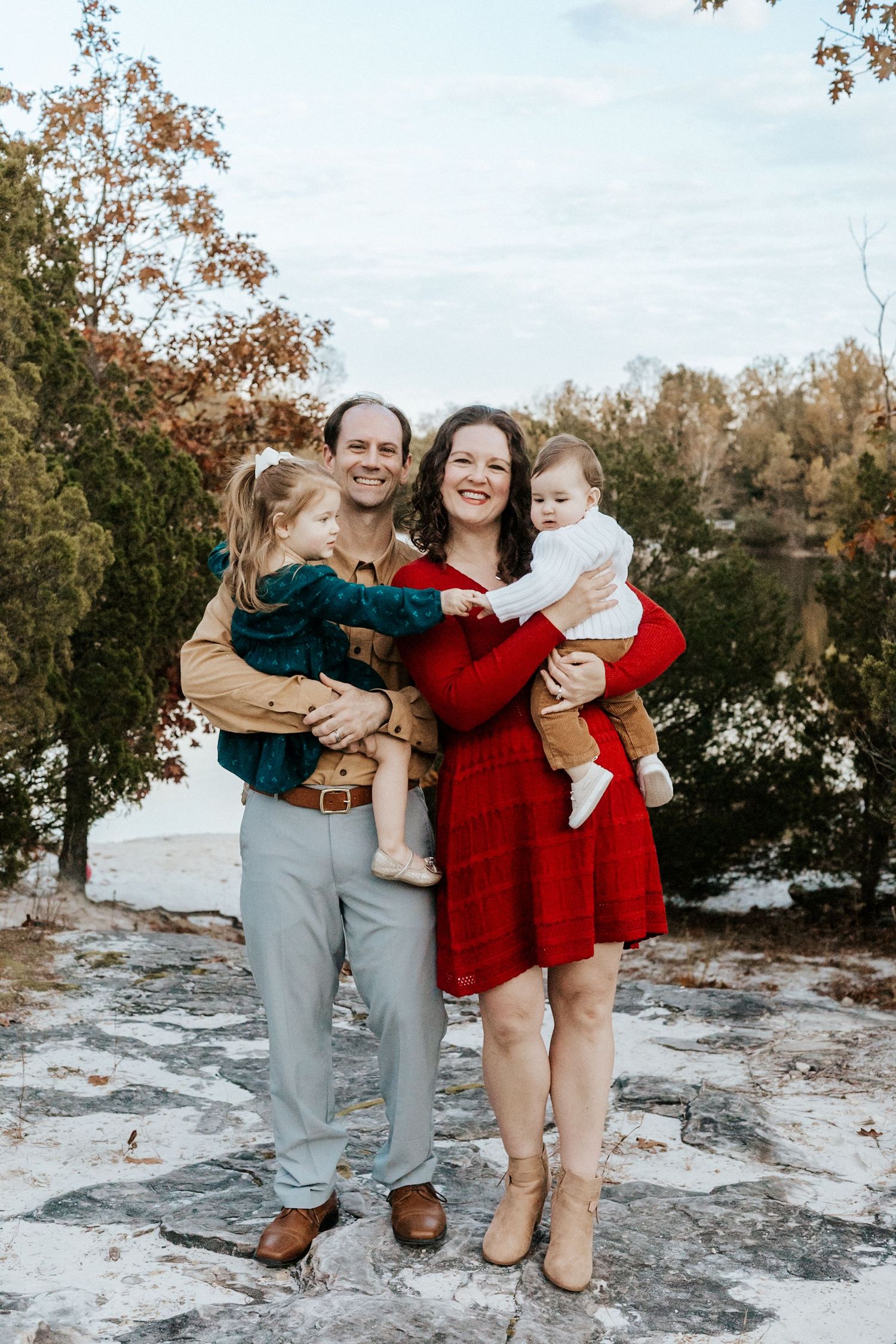 Happy family of four posing outdoors in fall, parents holding two young children, wearing festive red and teal outfits.