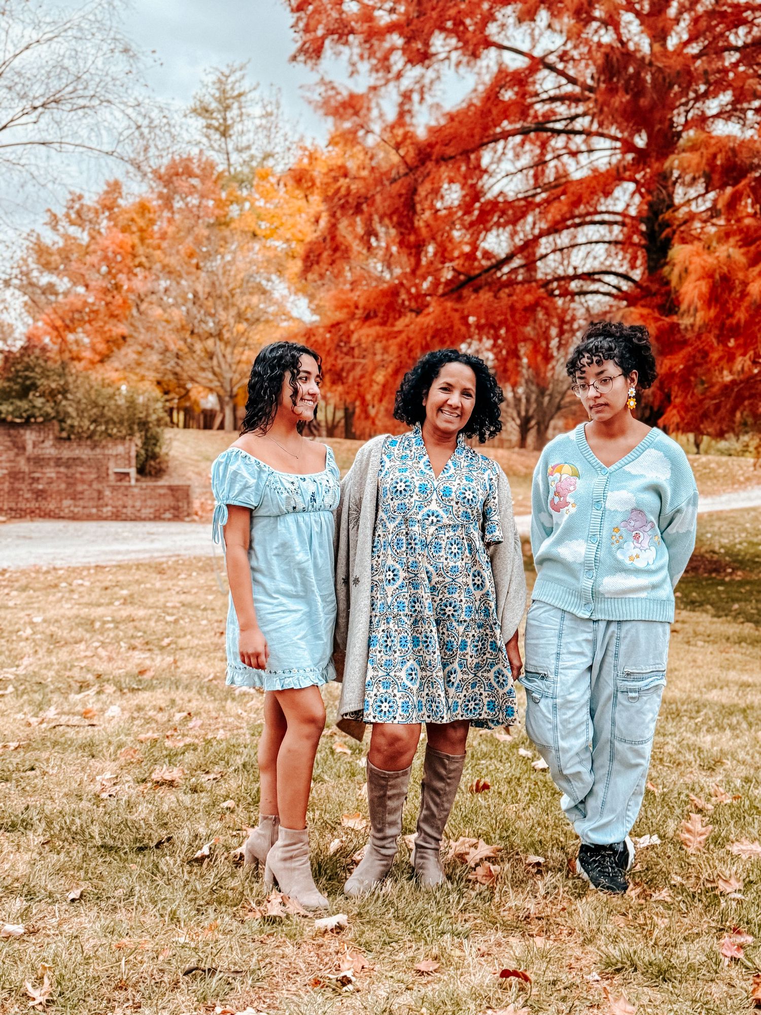 Three women in light blue outfits pose outdoors among vibrant red and orange autumn trees on a sunny fall day.