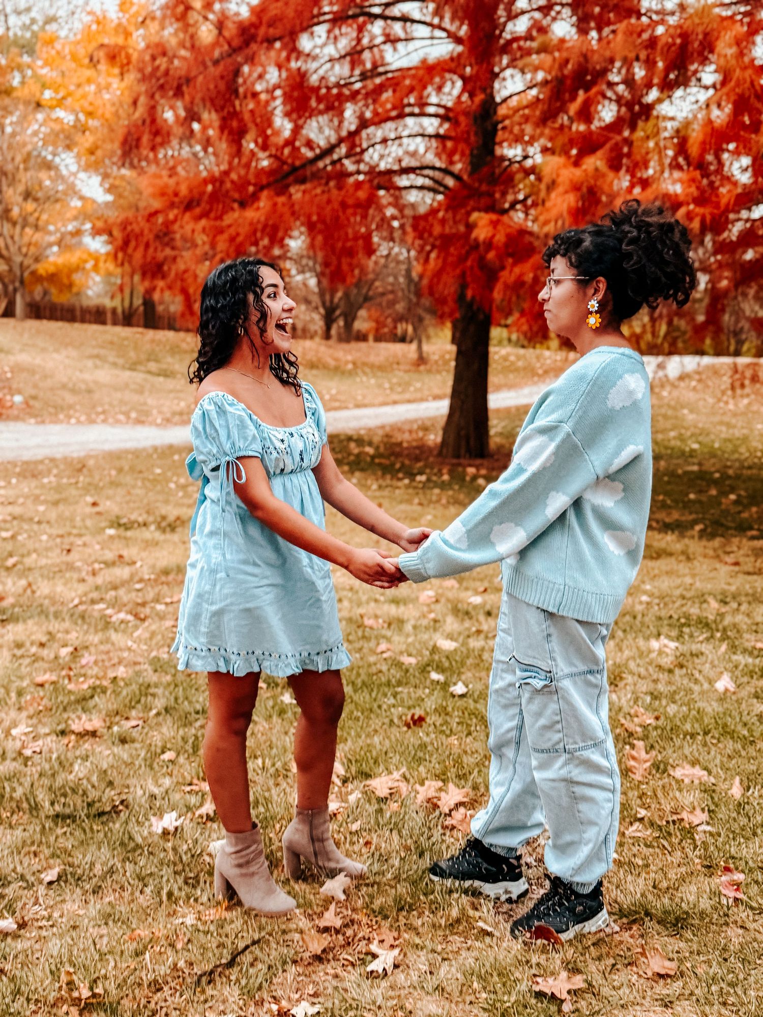 Two women in matching light blue outfits holding hands in a park surrounded by vibrant fall foliage.