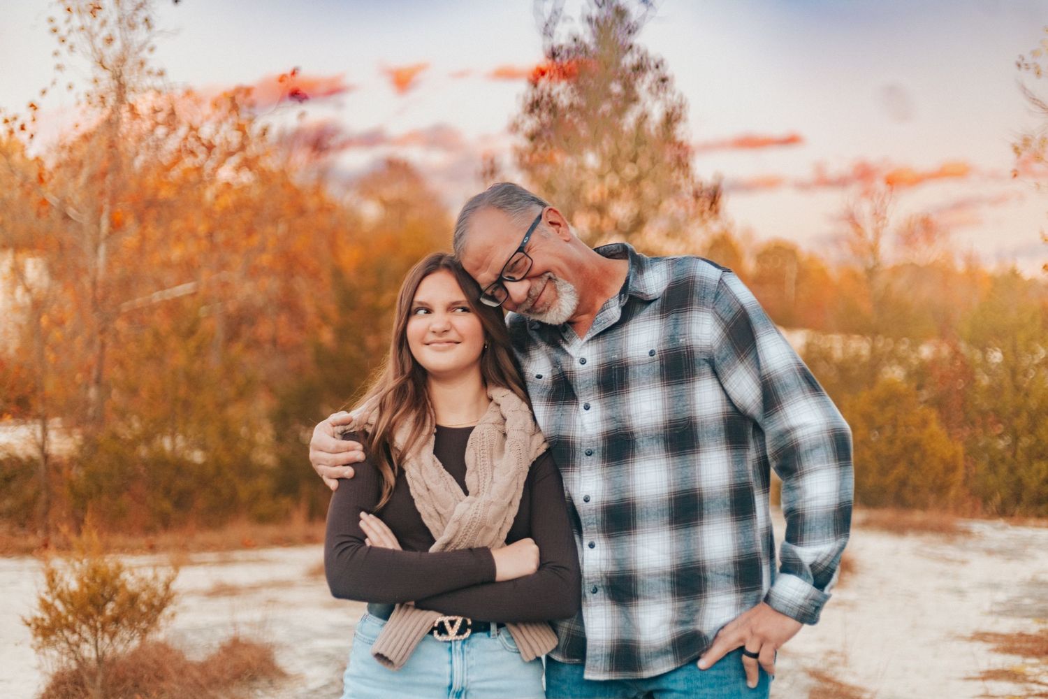 Couple smiling and laughing together outdoors during golden hour with autumn trees behind them.