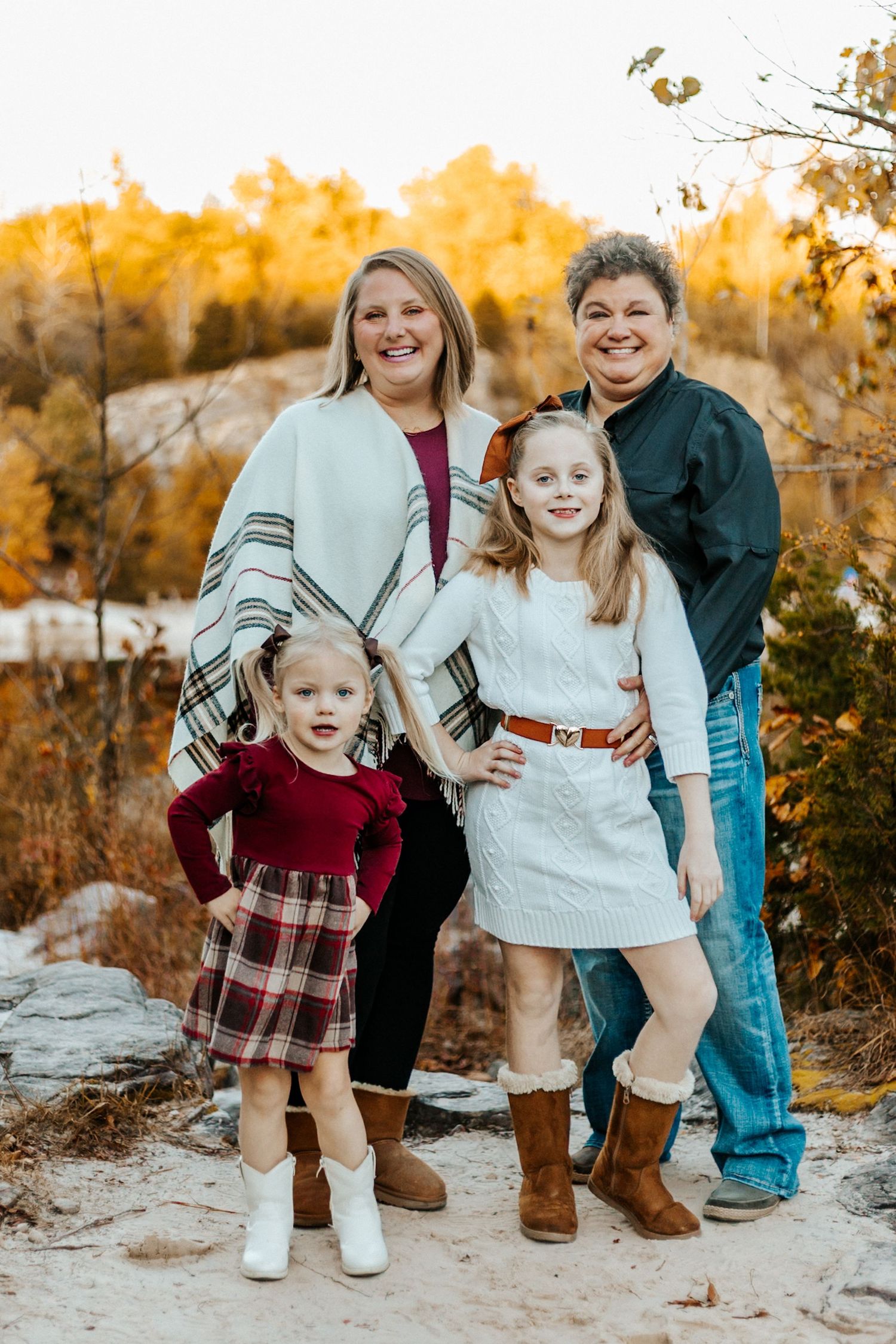Family of four posing outdoors in autumn, two women and two girls in festive attire, surrounded by fall foliage.