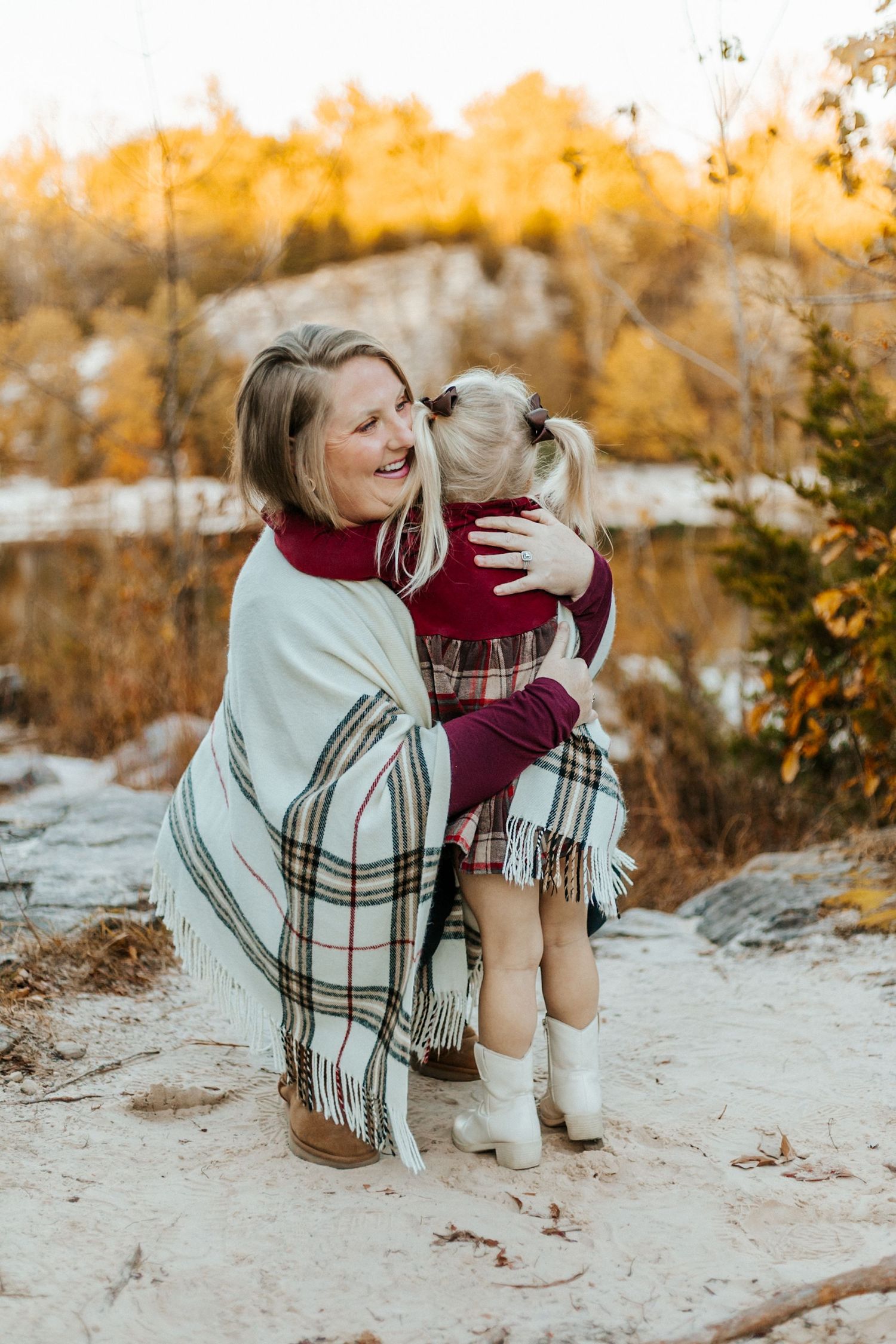 A smiling woman wrapped in a plaid blanket hugs a young girl in a maroon dress outdoors in autumn.