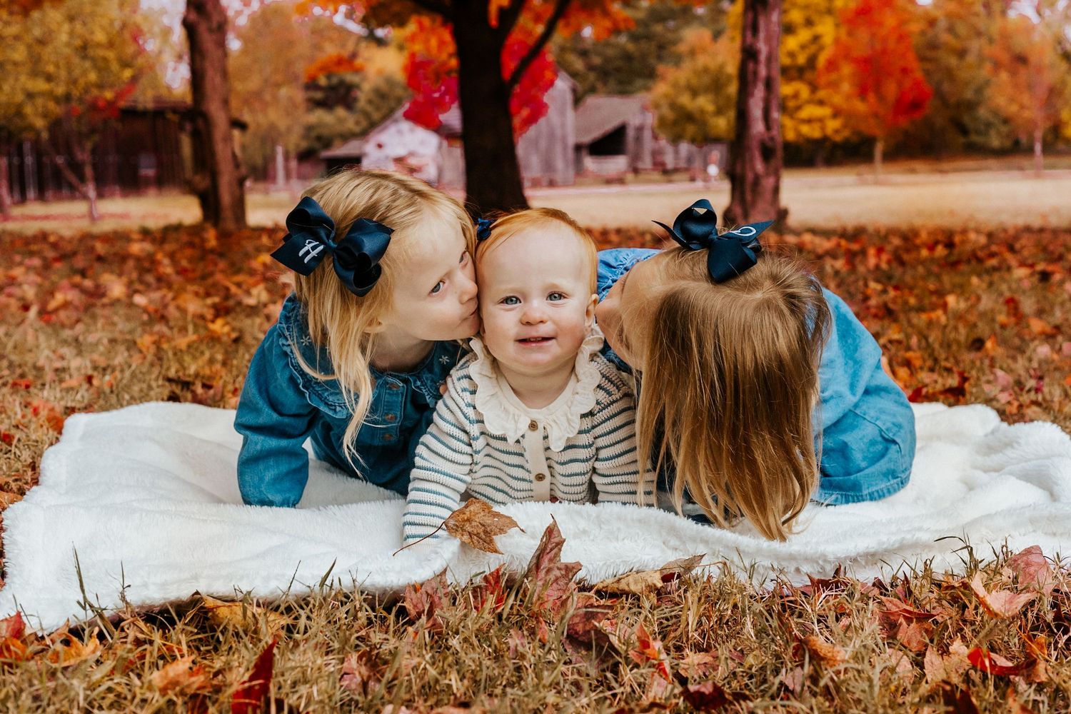 Two older sisters in denim jackets kiss a smiling baby on a white blanket surrounded by colorful fall leaves.