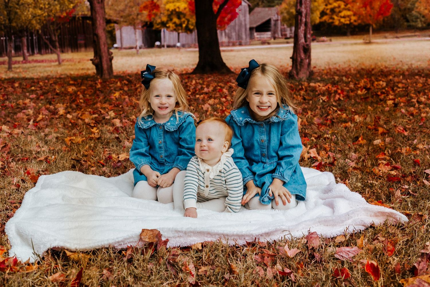 Three young siblings pose on a white blanket among autumn leaves, two girls in teal jackets and a baby boy smiling outdoors.