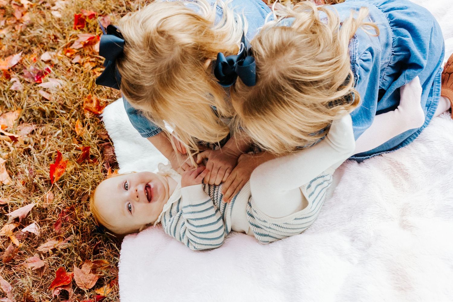 Two blonde girls in blue dresses lean over a smiling baby on a white blanket surrounded by fall leaves.