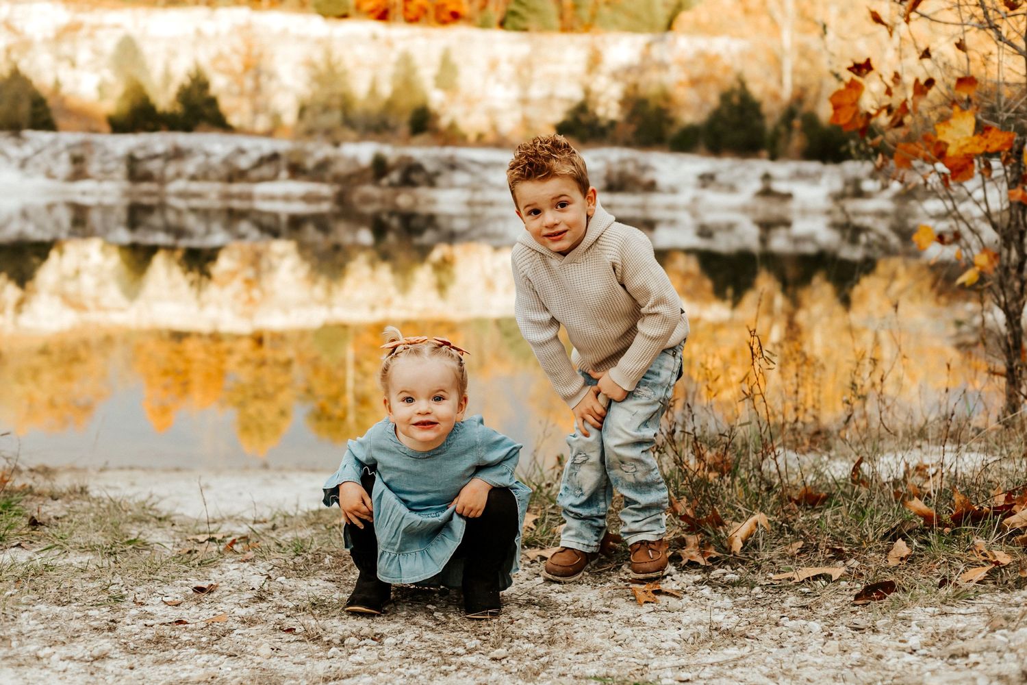 Two young children pose near a reflective autumn pond surrounded by colorful fall foliage.