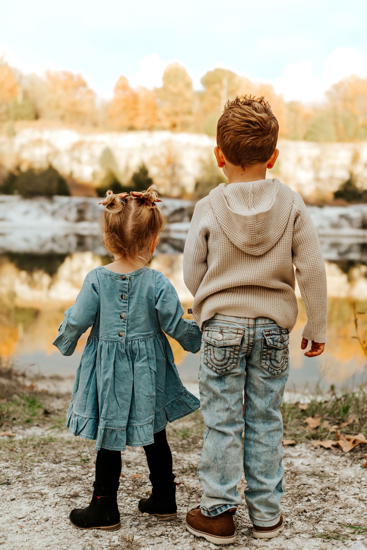 Two young children holding hands while gazing at a scenic autumn pond, girl in teal dress and boy in beige sweater and jeans.