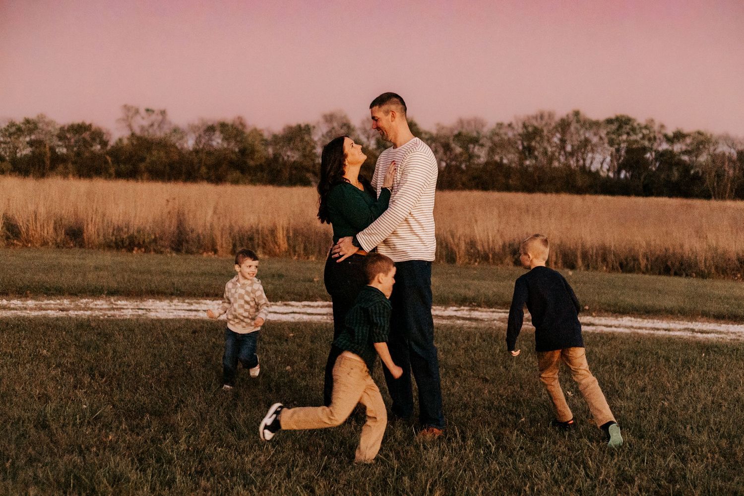 Family of four poses at golden hour in a field, parents embracing while two young boys play nearby.