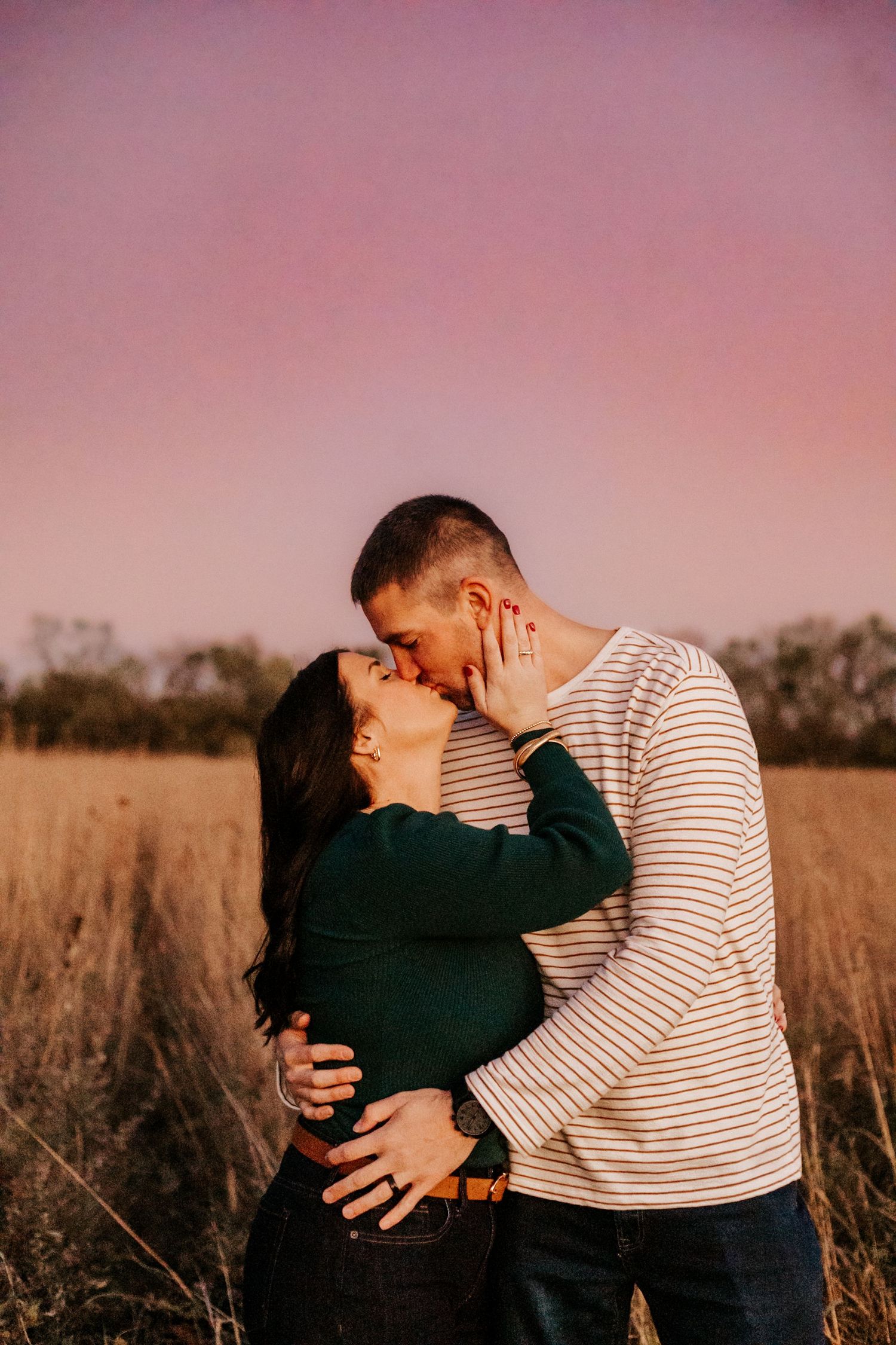 Couple sharing a romantic kiss in a golden field at sunset under a pink and purple sky.