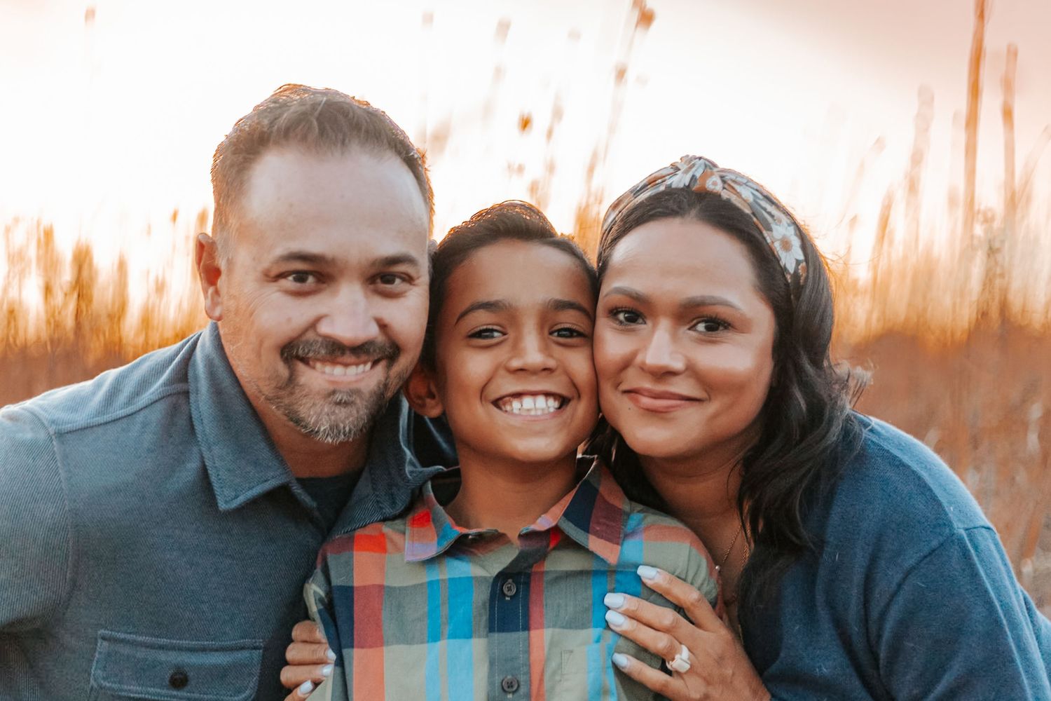 A happy family of three smiling outdoors at golden hour, with parents embracing their young son in a field.