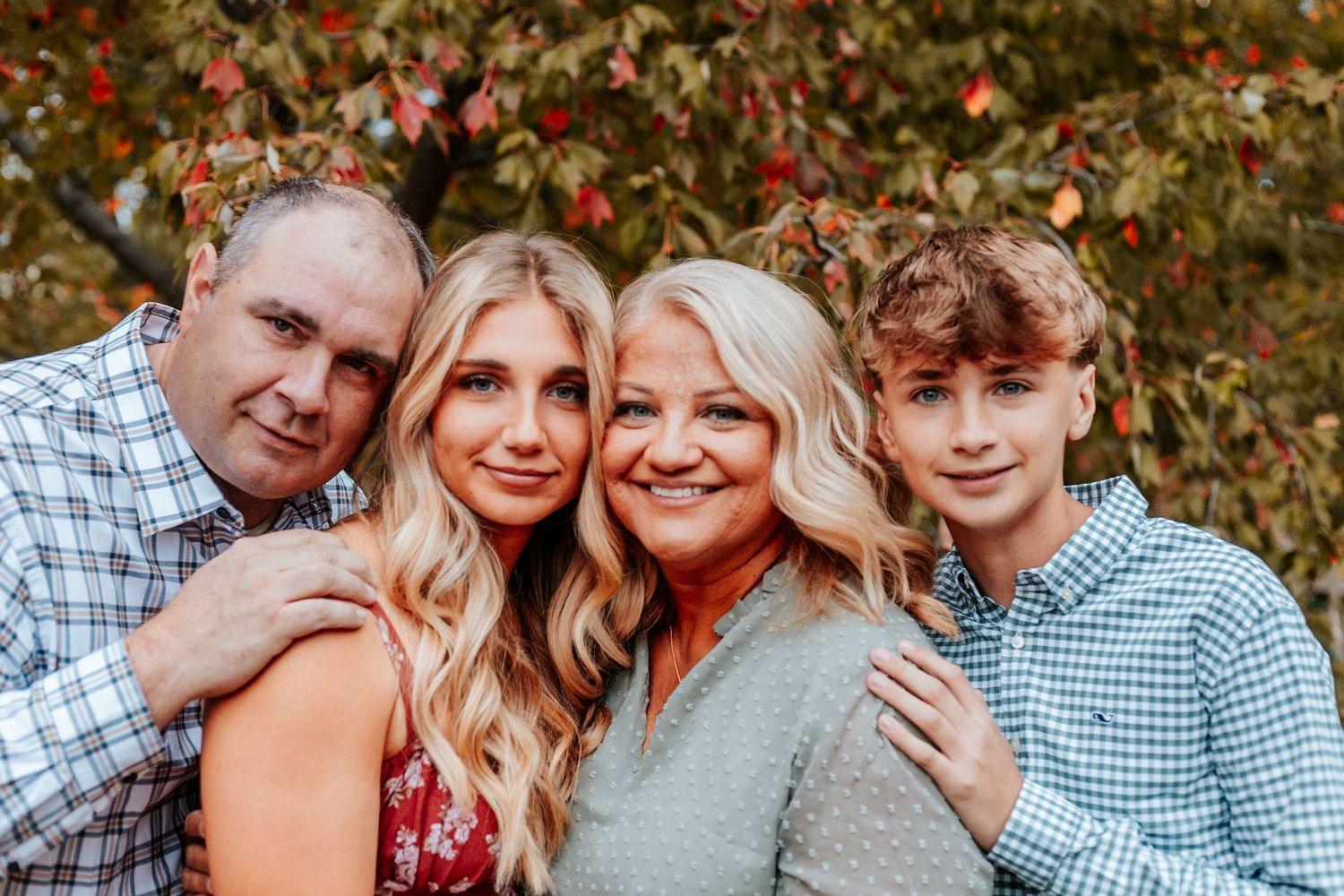A smiling family of four poses closely together outdoors with colorful autumn foliage in the background.
