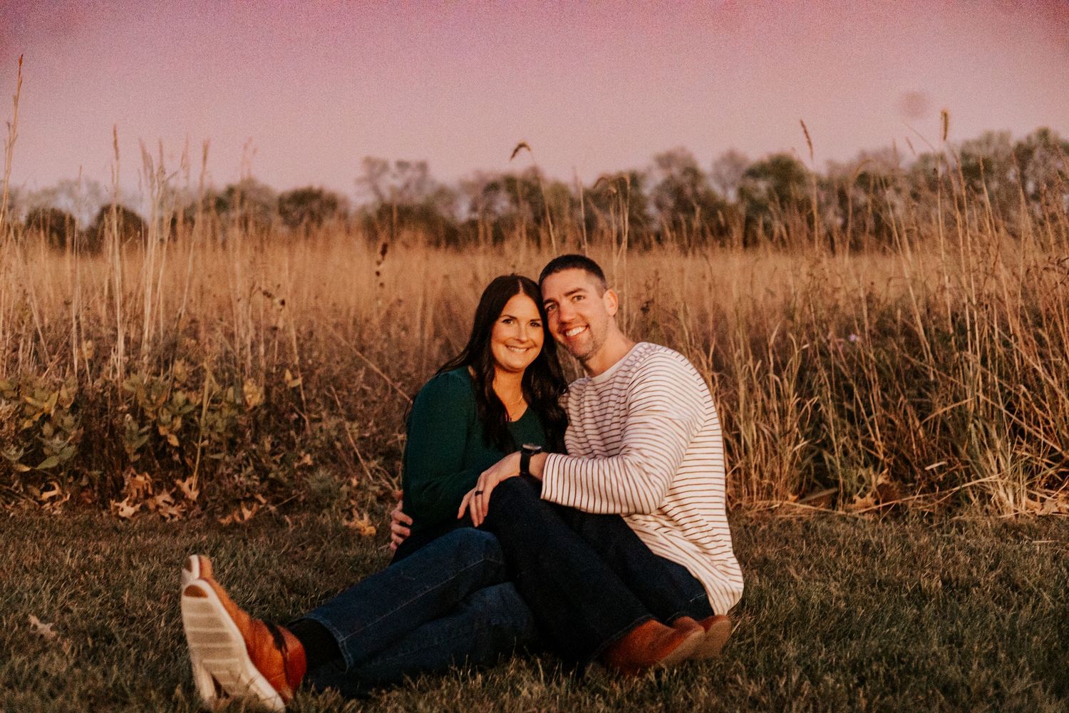 Couple sitting together in a golden field at sunset, smiling warmly during an outdoor engagement photo session.