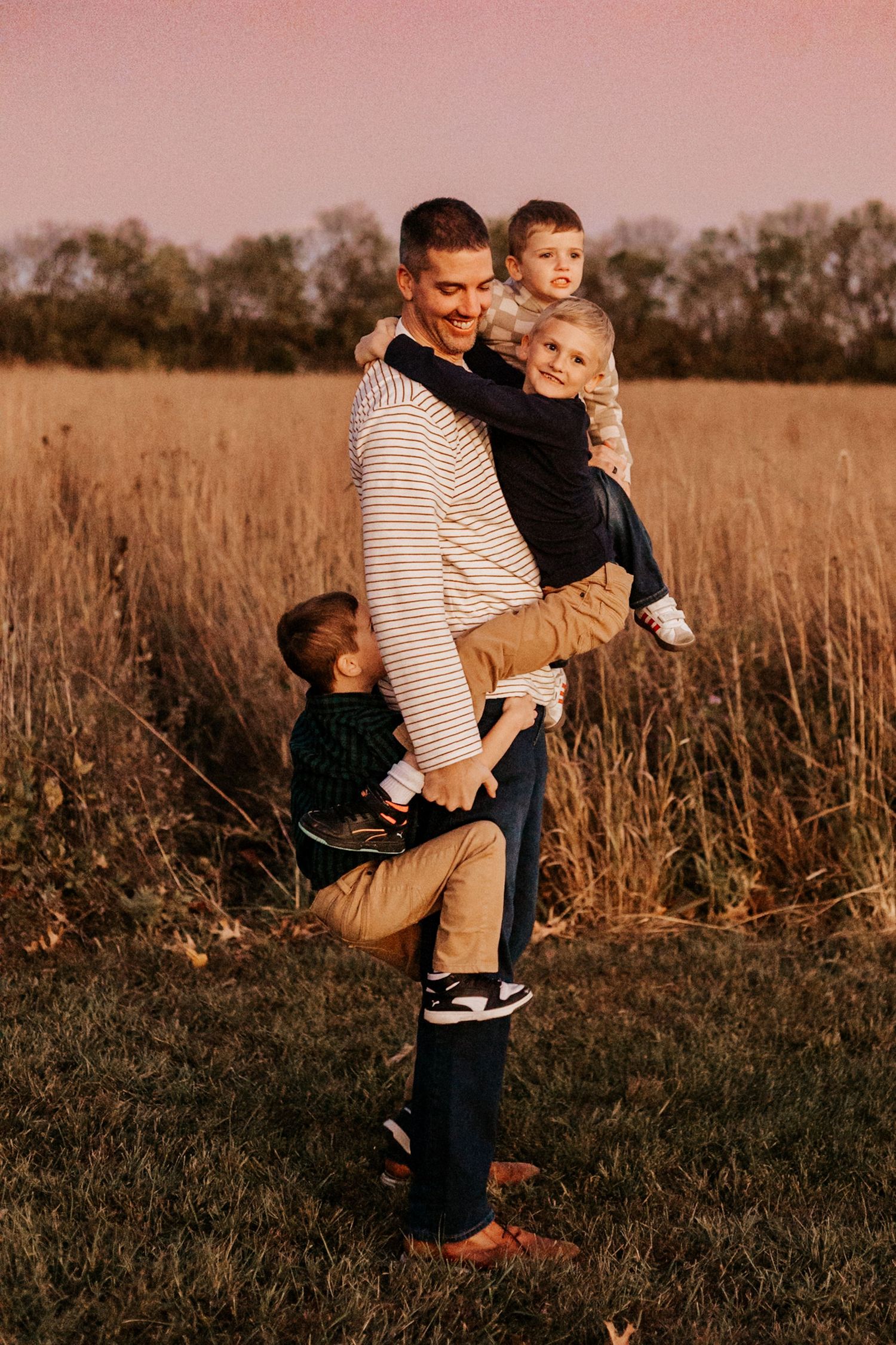 A father holds two young children in a golden field at sunset, while a third child clings to his leg.