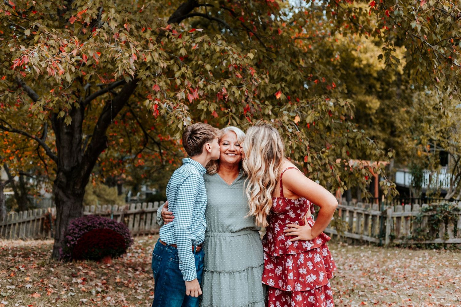 Two people kissing a smiling blonde woman on the cheeks outdoors among colorful autumn trees and fallen leaves.