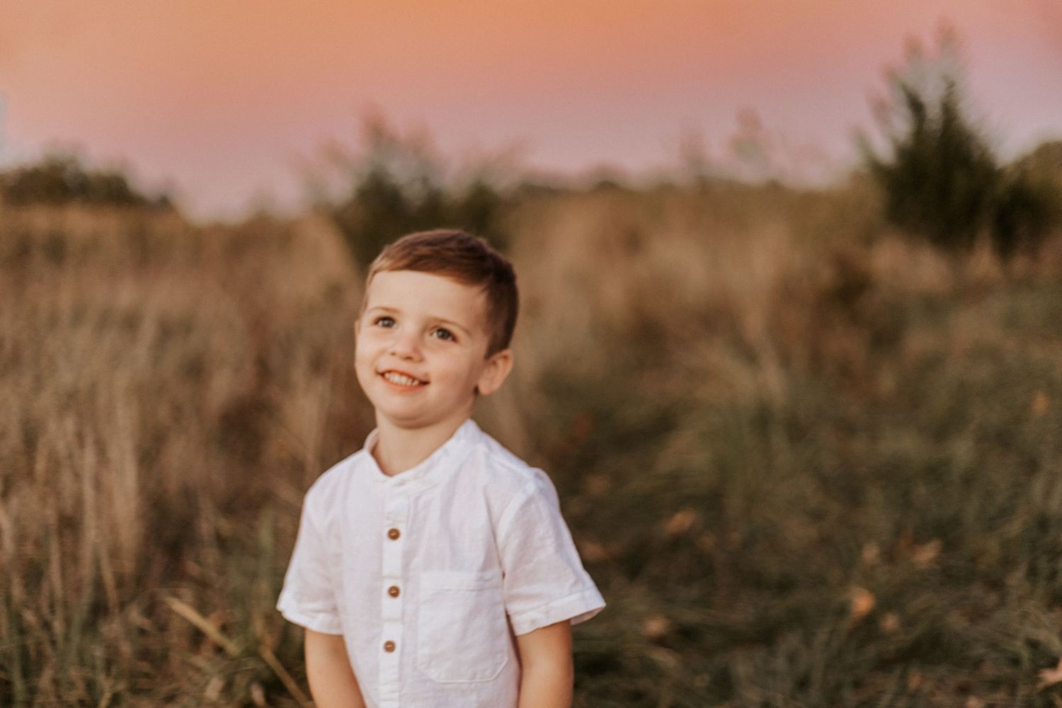 A young boy in a white shirt smiles in a golden field at sunset with a pink sky backdrop.