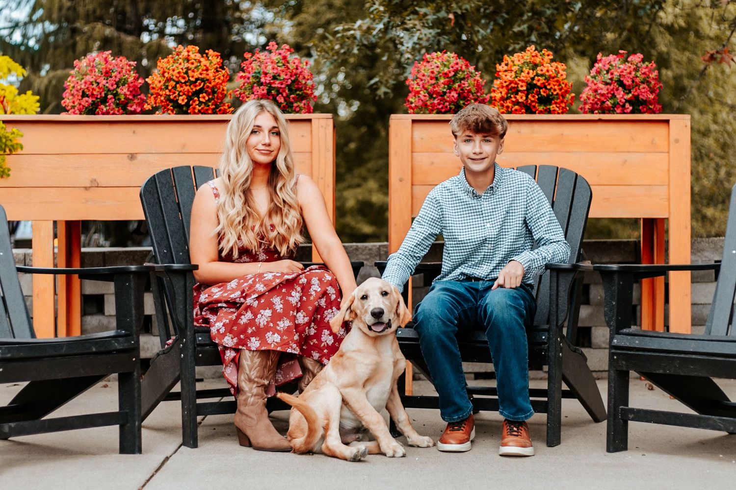 A young couple sits in black Adirondack chairs with a yellow Labrador dog, surrounded by colorful fall flower planters.