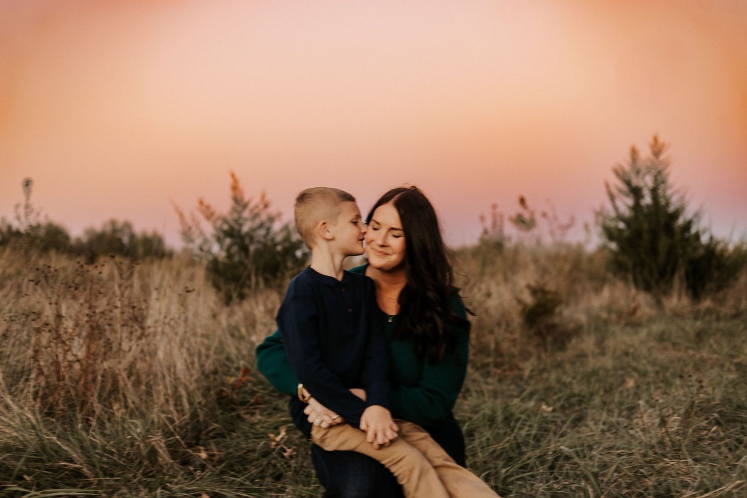 A mother holds her young son in a golden field at sunset, sharing a tender moment with a pink sky backdrop.