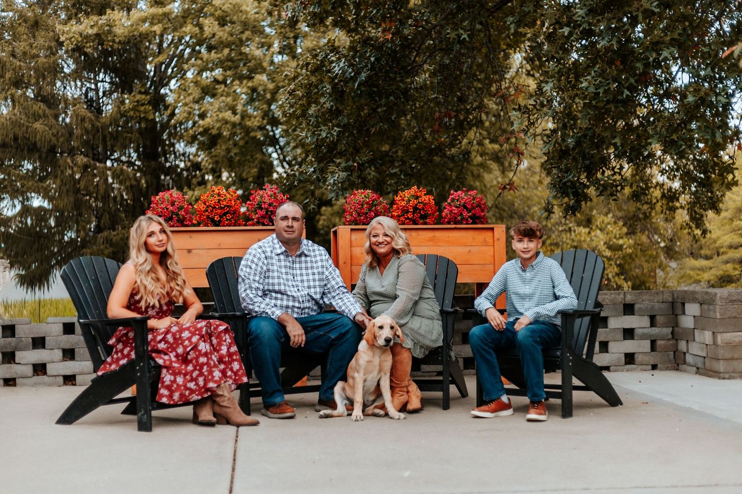 A family of four with a golden dog sits on black chairs on a patio with red flowers behind them.