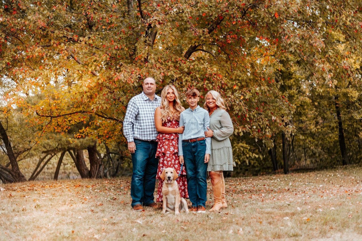 Family of four posing with a yellow dog among vibrant autumn trees in a fall outdoor portrait.