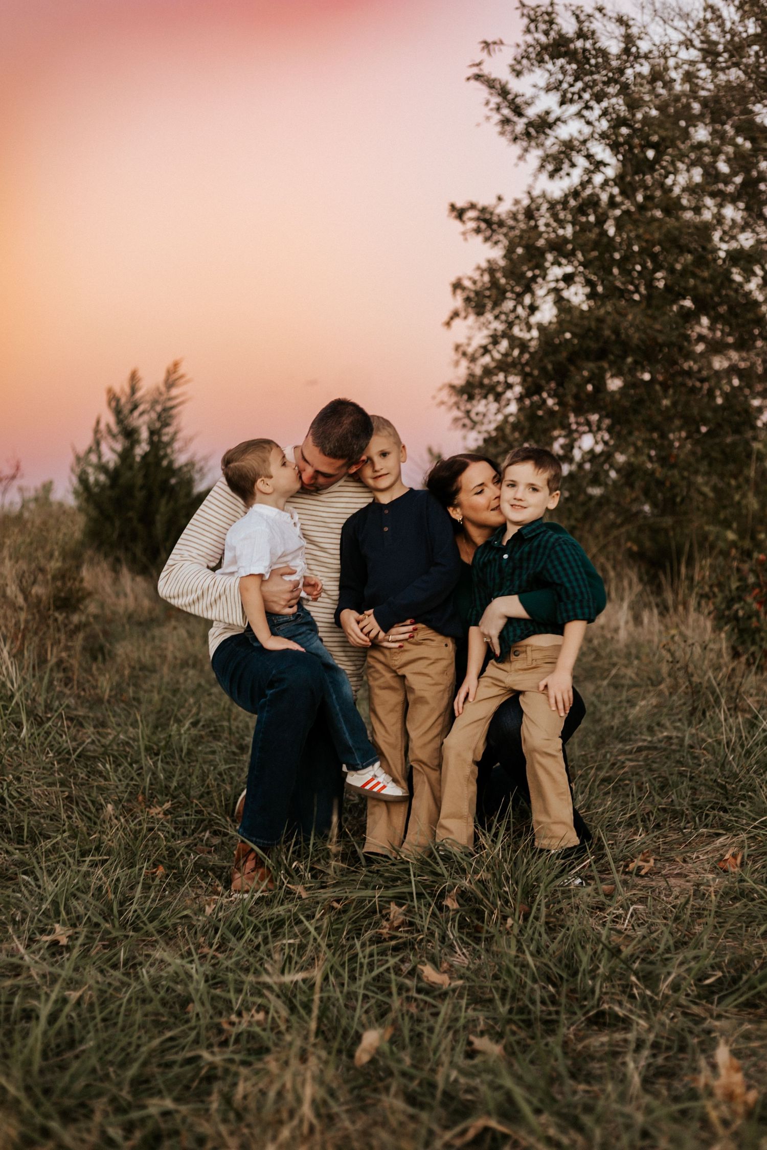 Family of five sharing a warm embrace in a golden field at sunset, parents cuddling three young boys outdoors.