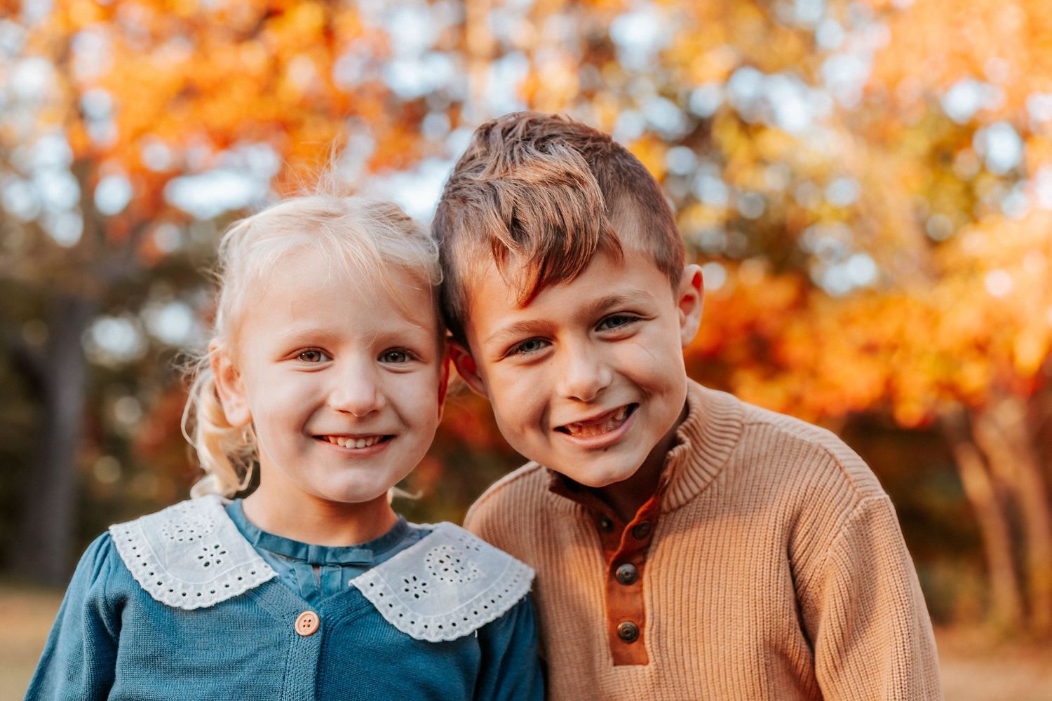Two smiling children pose together outdoors among vibrant autumn orange foliage in a warm fall portrait.