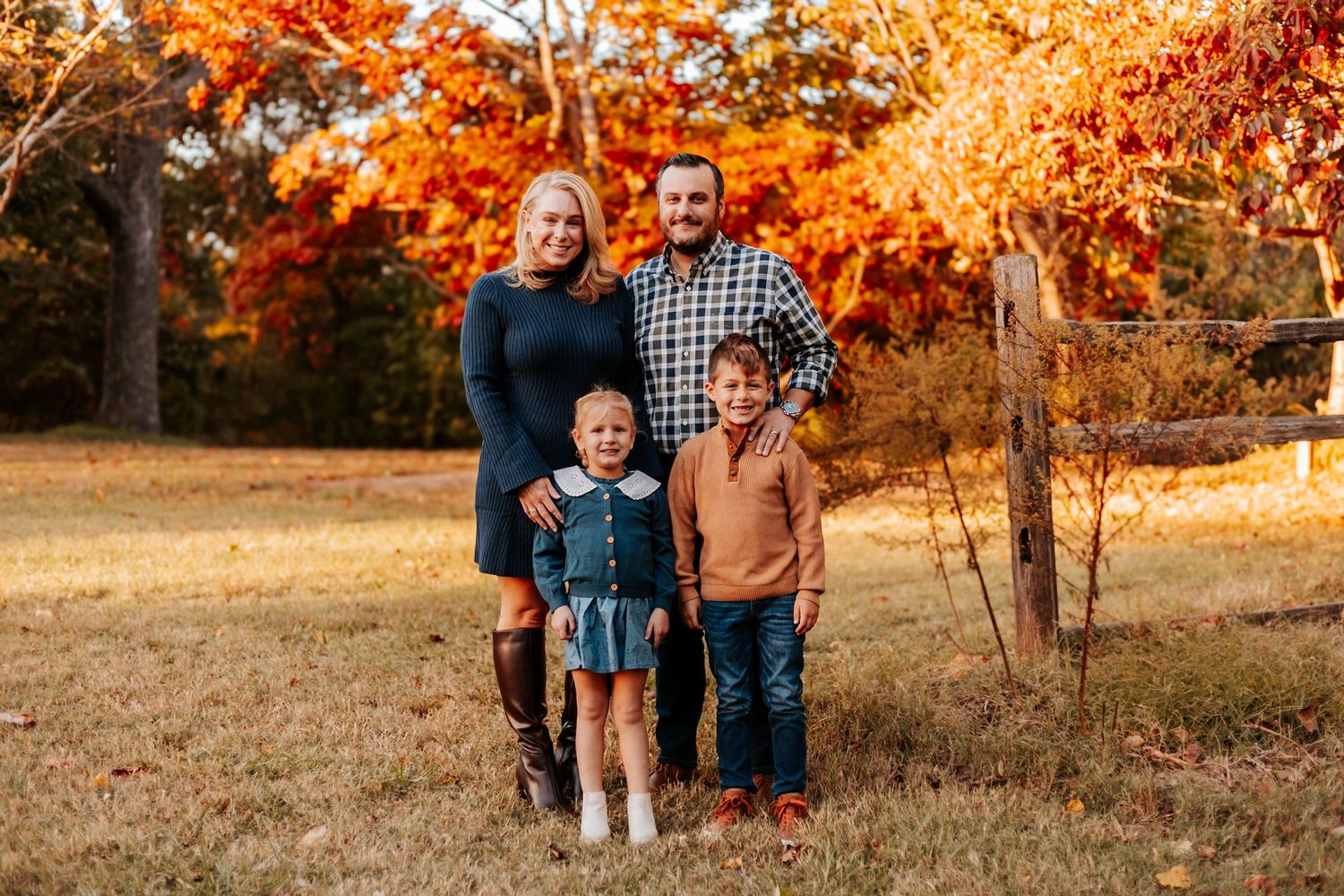 Family of four posing outdoors in fall with vibrant orange and red foliage in the background.