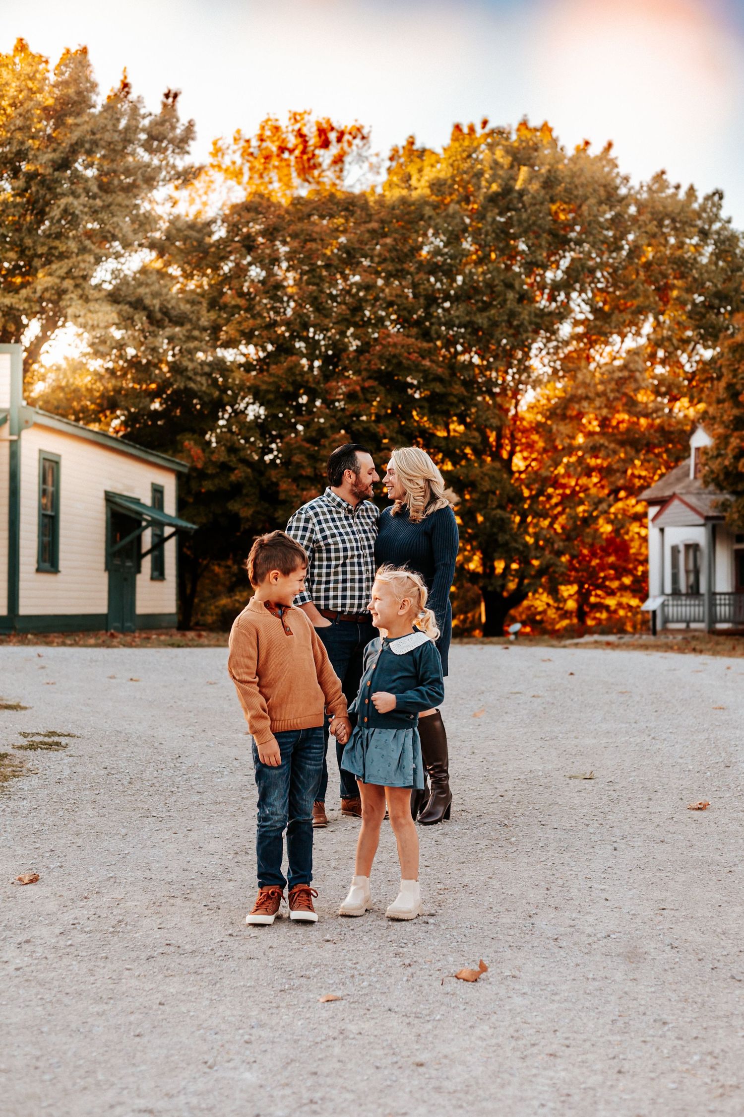 Family of four poses outdoors in autumn, parents gazing at each other while two young children stand in foreground on gravel path.
