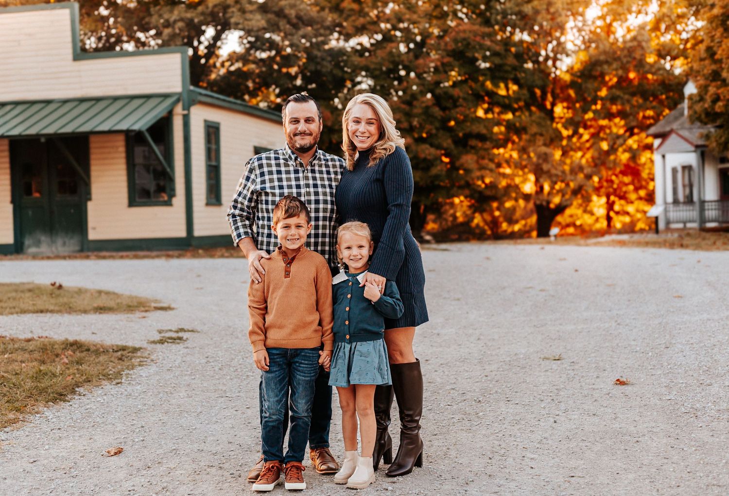 Family of four posing outdoors in fall foliage, parents and two children smiling in autumn attire.