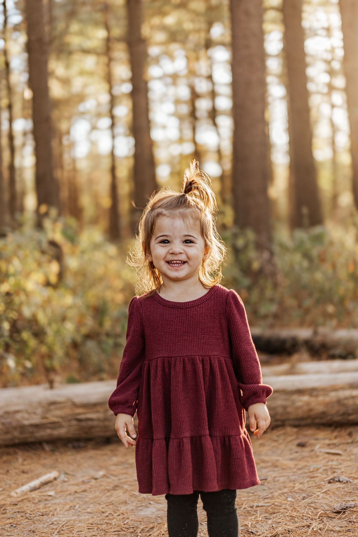 A smiling toddler girl in a burgundy dress stands in a sunlit forest during autumn.