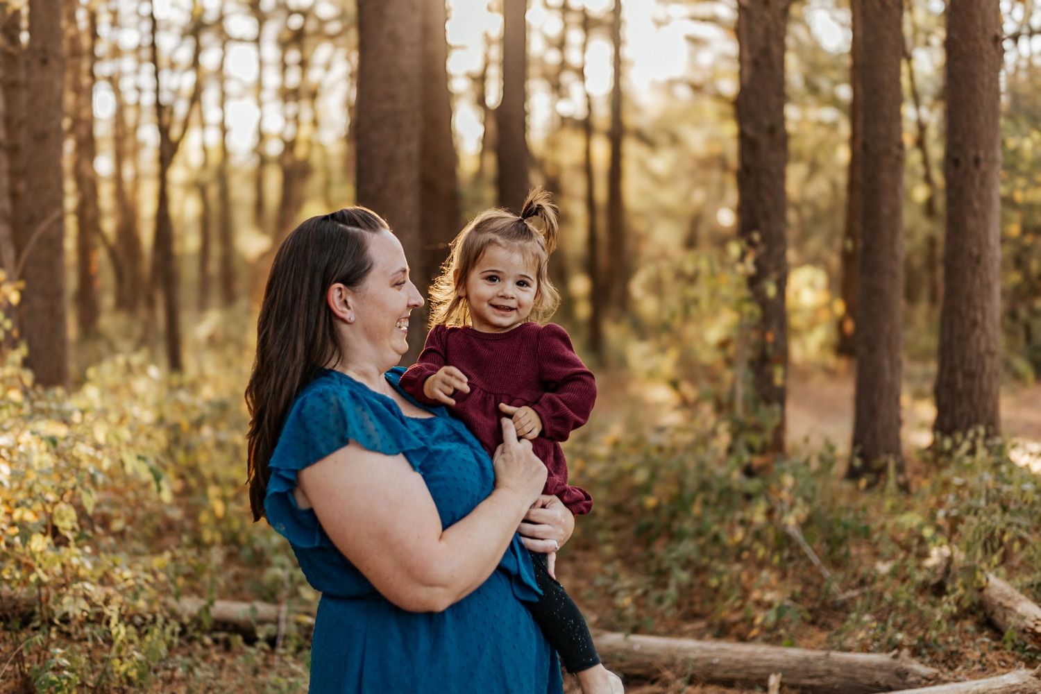 A mother holds her toddler in a sunlit forest, both smiling warmly in a golden autumn setting.