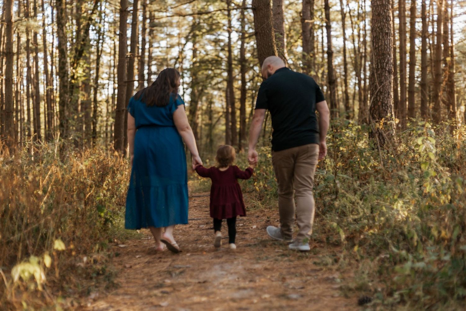Family of three walking hand-in-hand on a forest trail during golden hour, parents guiding young child in red dress.