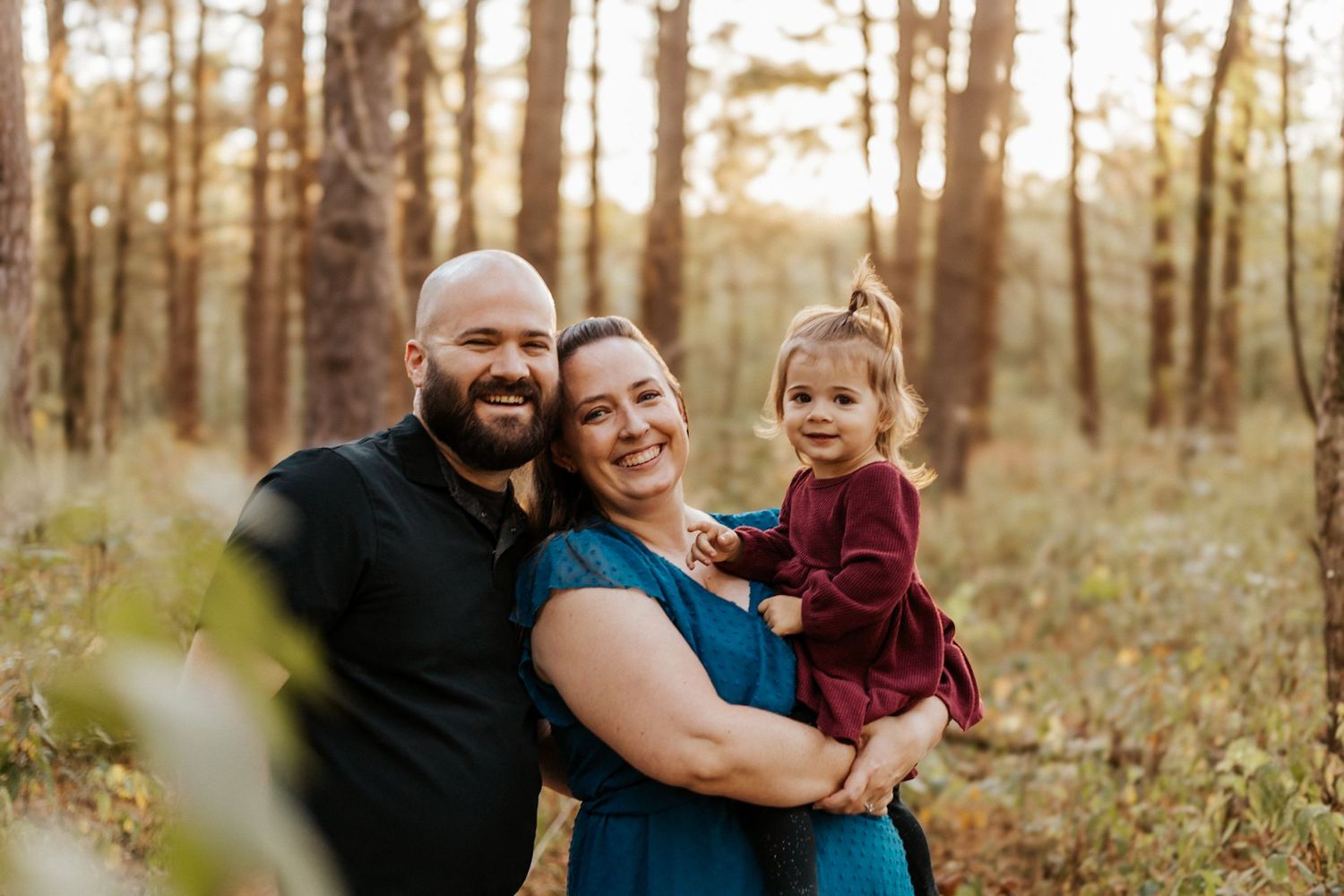A smiling family of three poses in a sunlit forest, with parents holding their toddler in a burgundy dress.